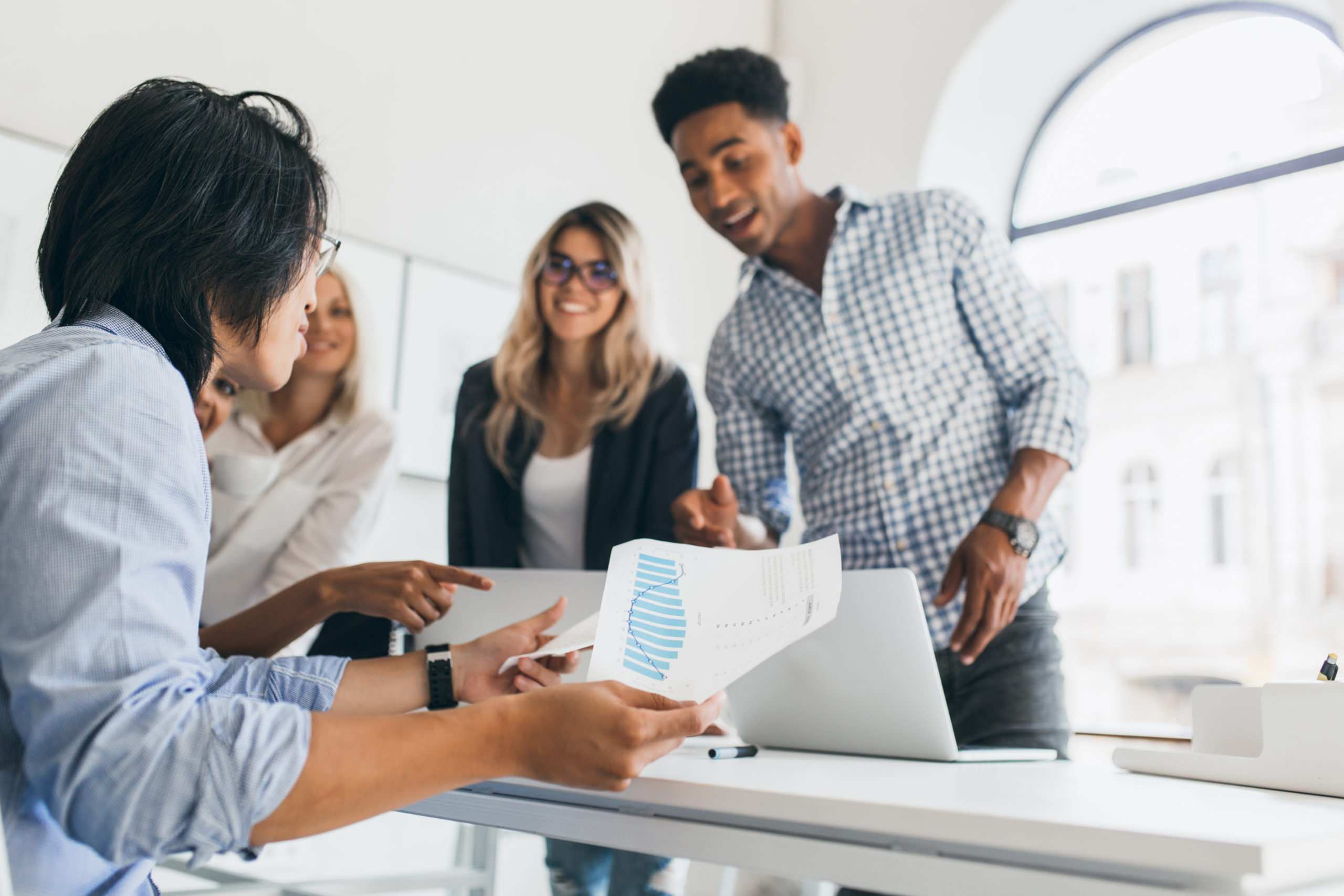 Asian manager in wristwatch sitting at his workplace and holding paper with graph. Indoor portrait of african programmer in checkered shirt talking with other employees in office..